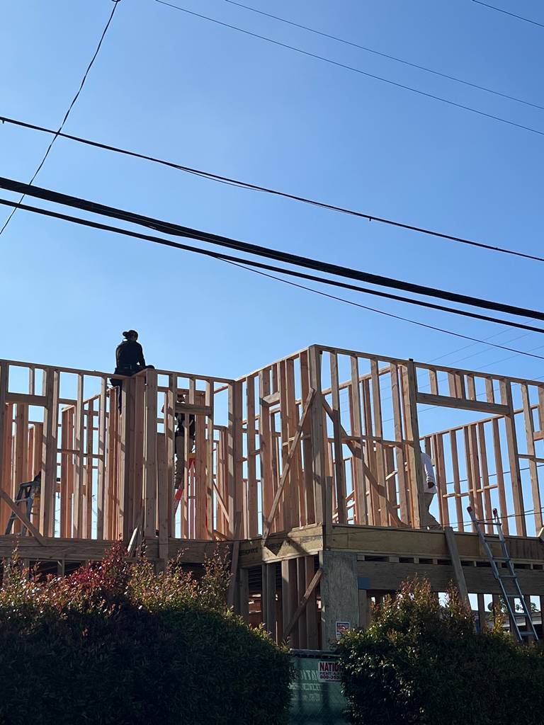 Framing of a second-story exterior wall is underway, with workers positioned on top assembling top plates and window headers.