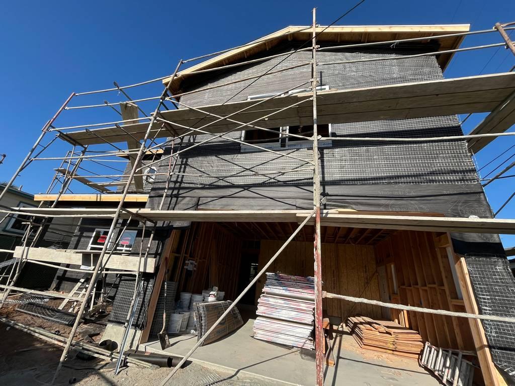 Frontal view of a house under construction with scaffolding, black lath wrap, and stacked stucco materials on-site.