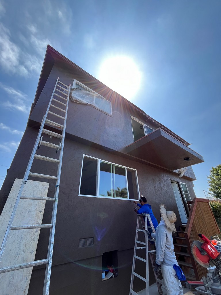 Workers accessing rear stucco wall with ladders, balcony, windows, and protected openings.